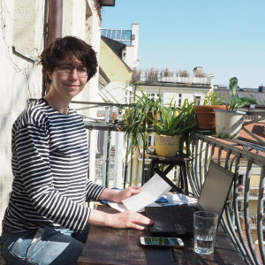 Foto im Hochformat von Rese am Schreibtisch vor dem Laptop sitzend und in Deine Richtung lächelnd. Der Arbeitsplatz ist auf einem Balkon im Sonnenschein über den Dächern von München.