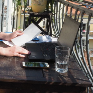 Fotoauschnitt: An einem sonnigen Ort steht ein Laptop auf dem Tisch, links Papier und recht ein Handy. Reses Hände bedienen den Arbeitsplatz.